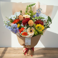 A large bouquet of fresh flowers including sunflowers, daisies, and delphiniums with a mix of other flowers in a variety of colors, wrapped in paper and tied with a ribbon.